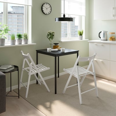 Small dining area with black table, white chairs, and green walls. Table set with cups and a vase. Chairs fold for space-saving.