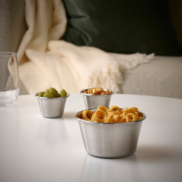 Three metallic bowls on a white table hold snacks: pretzels, mixed nuts, and green olives. Perfect for outdoor gatherings.