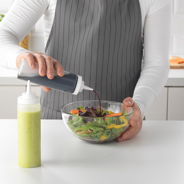 A person pours dressing from a grey squeeze bottle onto a salad in a bowl on a kitchen counter.
