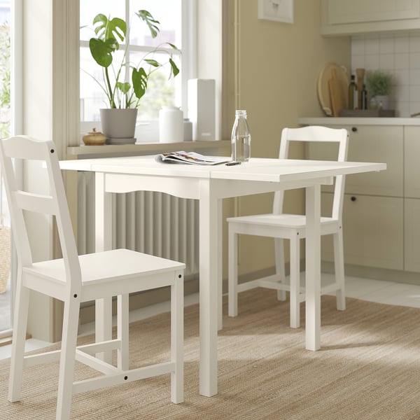 Small white wooden dining table with two chairs, placed near a window with a plant, set with a bottle and napkins.