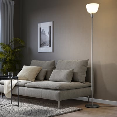 A modern living room with grey sectional, patterned pillows, glossy lamp, side table, large plant, and black-and-white wall art.