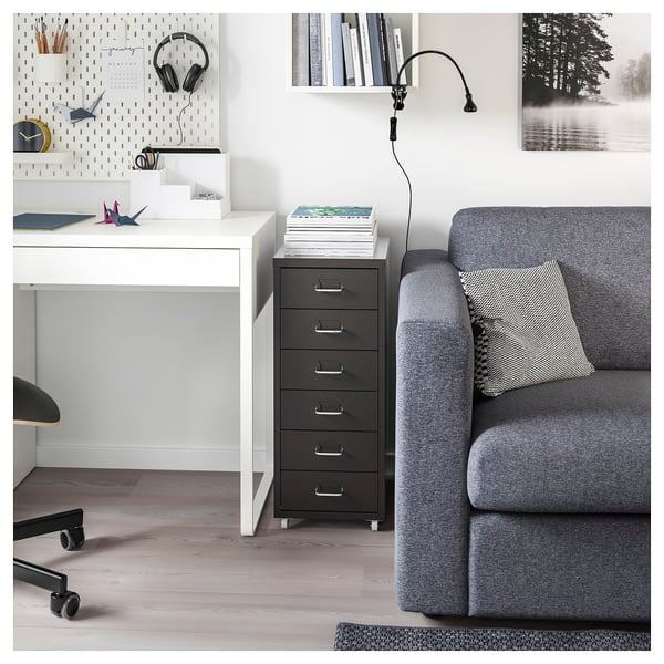 A HELMER black metal filing cabinet with five drawers on wheels beside a white desk and a grey armchair with a throw pillow.