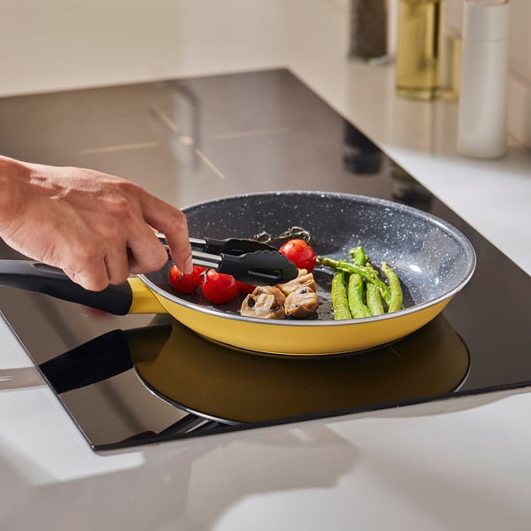 Hand tosses ingredients in a yellow, non-stick frying pan on a stovetop, showcasing even heat distribution and easy lifting with its ergonomic handle.