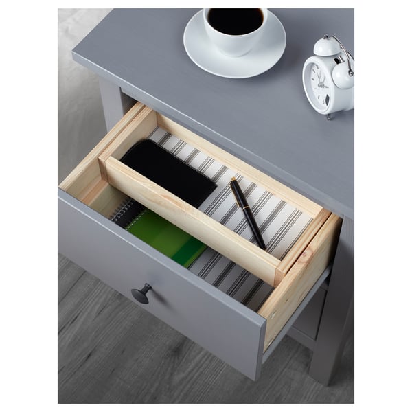 Grey bedside table with open drawer revealing pens and notebook in a wooden organiser, alarm clock, and coffee on top.