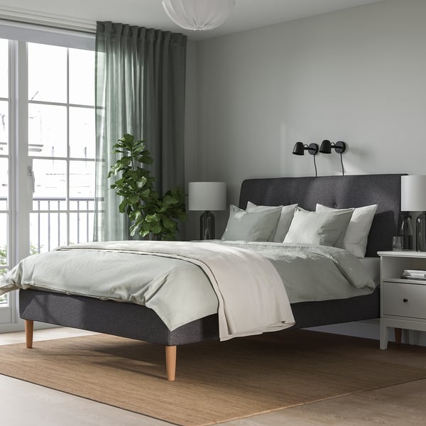 Modern bedroom with upholstered IDANÄS bed, dark grey, sloping headboard, wooden legs. Minimalist design with neutral tones, large window, rug, plant.