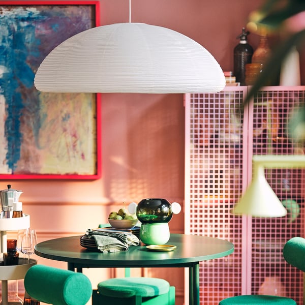 Large white rice paper pendant lamp hangs prominently over small table with green stools. Bright colour wall and pink partition in background.
