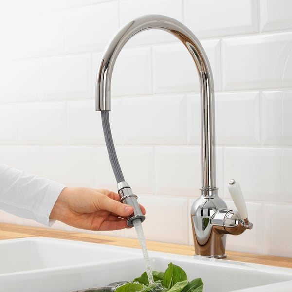 A person turns on a chrome kitchen tap, releasing water into a sink. The high, pull-out spout is functional for rinsing.
