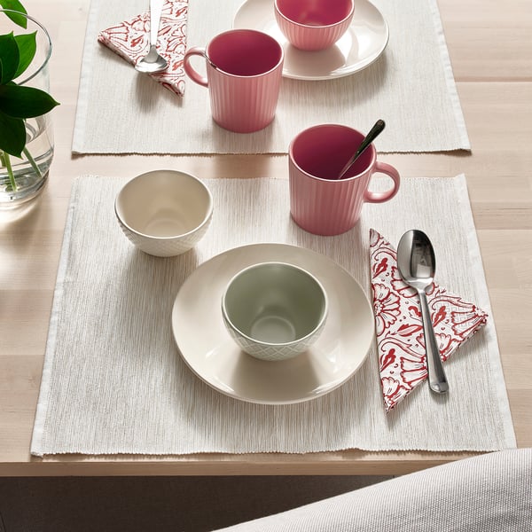 Table set with pink mugs, green bowl, white plate, spoons, and patterned napkins on beige placemats.