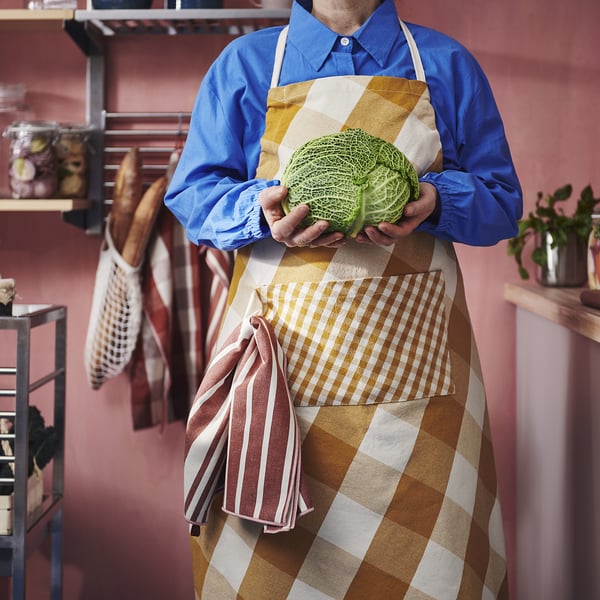 Person in KÖSSEBÄR chequered apron holding greens, kitchen backdrop.