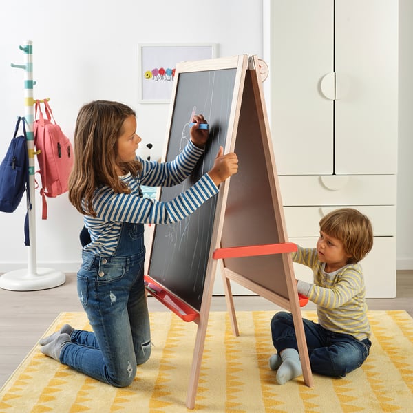 Two kids use a foldable MÅLA easel, one drawing on a chalkboard, the other observing.