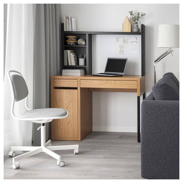 Modern home office setup with MICKE desk, white swivel chair, grey curtains, and blue sofa. Wooden desk with drawers, laptop, books, and lamp.