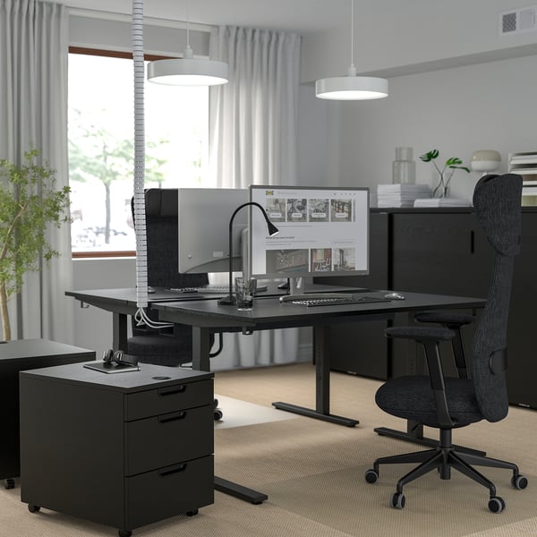 Modern home office with height-adjustable desk, chair, monitor, lamp, and plants. Large window and stylish lighting.