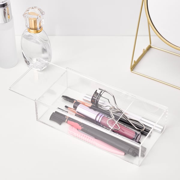 Clear acrylic organiser box on a white surface, holding makeup tools like eyelash curler and tweezers. Gold-framed mirror is visible to the side.
