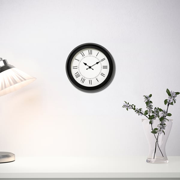 Modern quartz clock with black frame hangs above table with lamp and flowers.