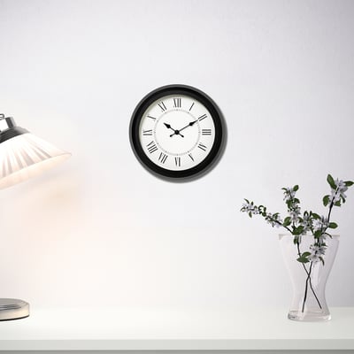 Modern quartz clock with black frame hangs above table with lamp and flowers.