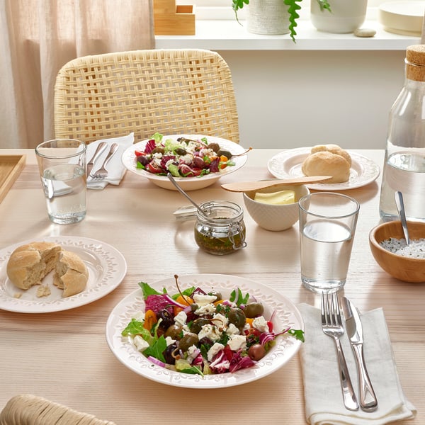 A wooden table set with white rimmed plates, colourful salads, bread rolls, glasses, and cutlery.