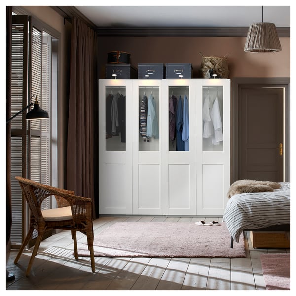 Modern bedroom white PAX wardrobe with glass doors displaying clothes. Accessorised with wicker chair, rug, and pendant lamp.