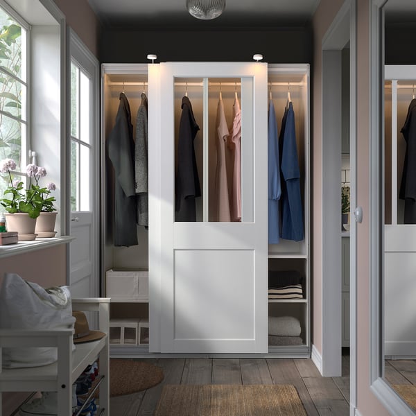 White PAX wardrobe with glass doors showing clothes and towels, beside a window with a plant.