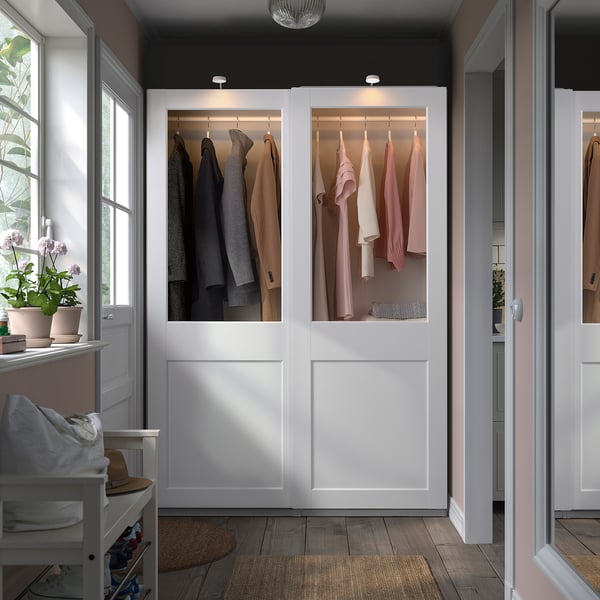 A white PAX wardrobe with glass doors displaying colourful clothes. It stands between windows and another wardrobe, offering ample storage.
