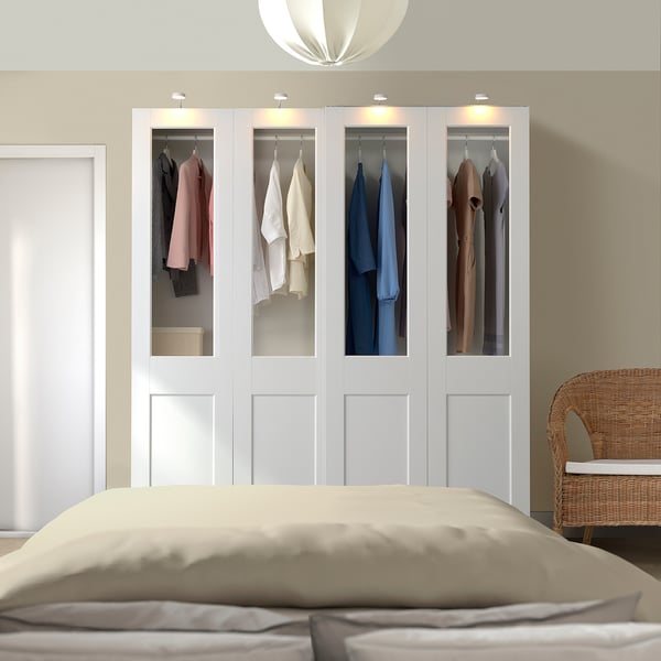 Modern bedroom with large PAX wardrobe and glass doors showing clothes, beside a bed with neutral linens and a wicker chair.