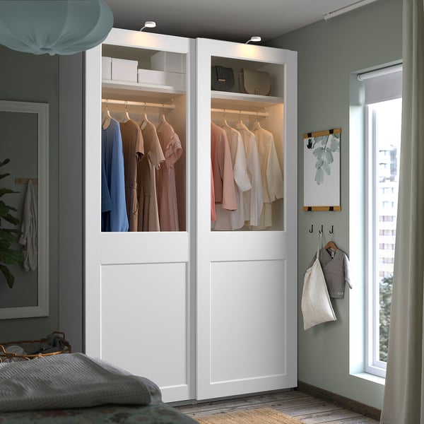 Modern bedroom featuring a large, white PAX wardrobe with glass doors showcasing clothes, situated next to a large window with curtains.