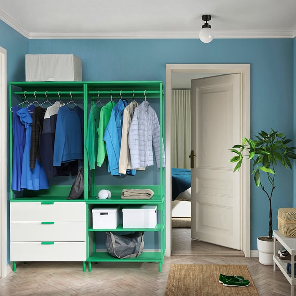 A bright green PLATSA wardrobe stands against a blue wall, housing clothes and storage bins, complementing a white door and plant nearby.