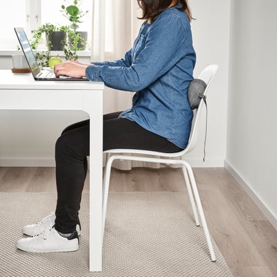 Person sitting at desk using laptop, wearing denim shirt, black pants, and white trainers, with a white chair cushion providing lumbar support.