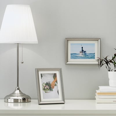 A silver picture frame on a table displaying a bouquet, near a lamp. The frame can stand or hang.