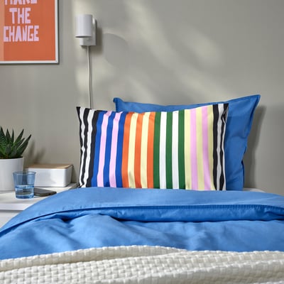 Bright blue SNÖDROTTNING pillowcase with stripes on a white bed, with a small plant, water glass, and poster.