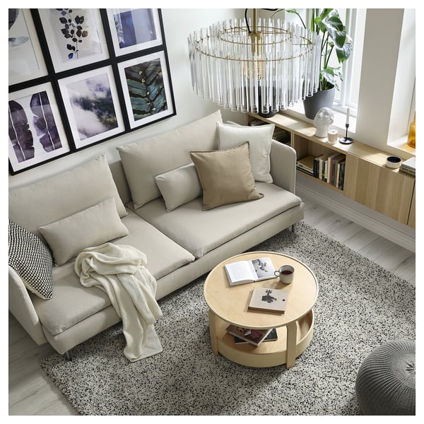 Modern living room a beige SÖDERHAMN sofa with pillows, a round wooden coffee table with books and a cup, framed wall art, and a stylish chandelier.