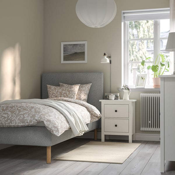 Cosy bedroom with grey TÄRNKULLEN bed, padded headboard, and wooden legs. Neutral bedding, bedside table with lamp, and large window with plant.