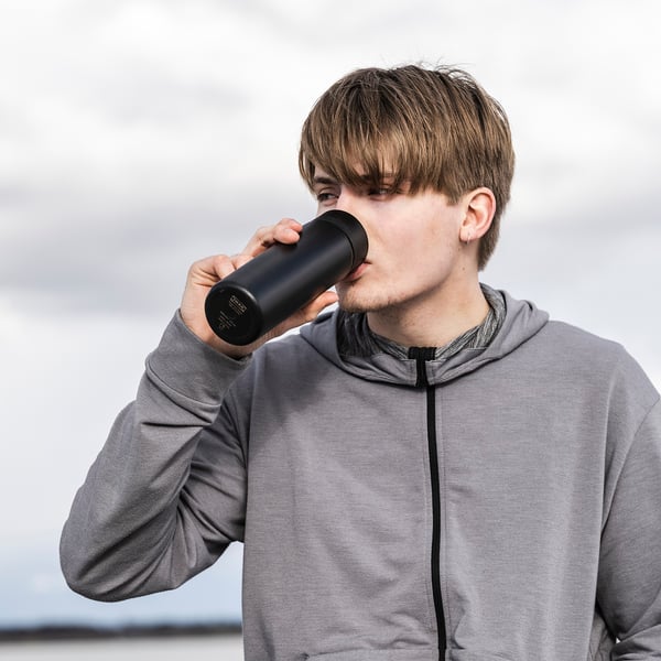 Young person drinks from black, cylindrical UNDERSÖKA travel mug with a grey hoodie.