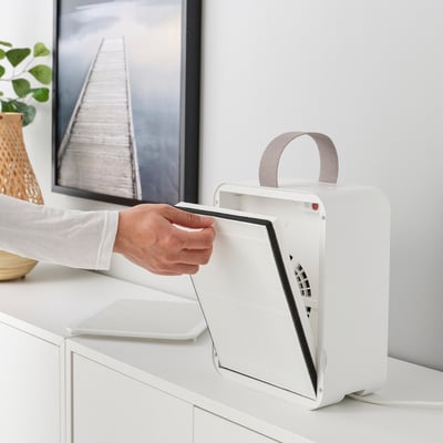 In a hand operates an air purifier, likely changing its philtre. The purifier has a detachable philtre, indicated by the handle and users action.