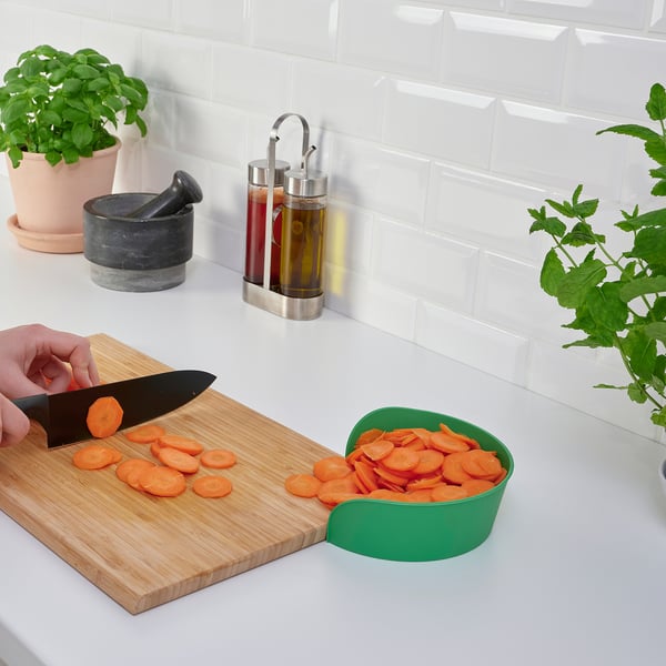 Person chops carrots on a wooden board, using a green bowl to collect slices, on a white counter with potted herbs and oil dispensers.