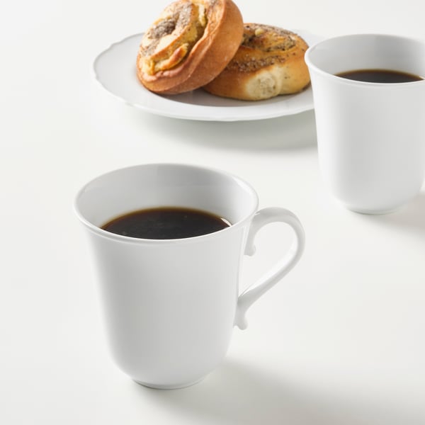 Two white mugs with coffee, one with handle, and pastry on plate. Mugs are durable, impact-resistant porcelain.
