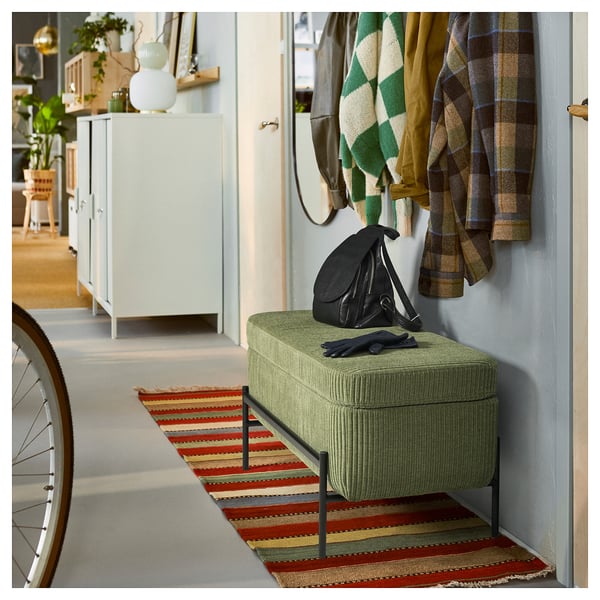 A green VALTORP storage bench in a hallway. It has a rectangular shape with raised edges and a cushioned seat. A black bag and gloves are on top.