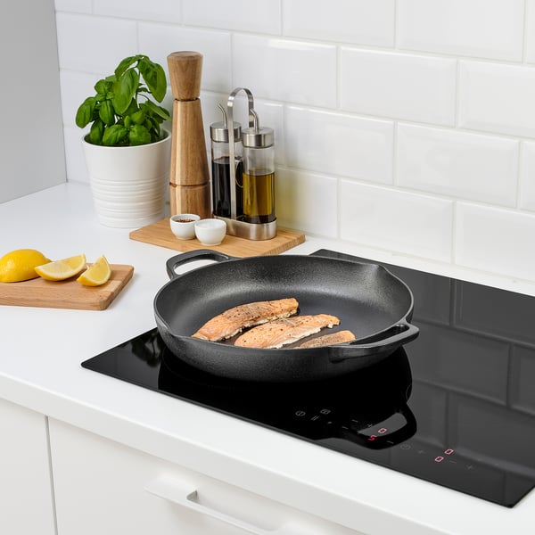 Modern kitchen with cast iron pan cooking fish on an induction hob. Lemon slices and fresh herbs nearby, set against white tiles.