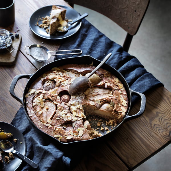 Cast iron skillet with cake on wooden table. Plate with cake slice, spoon, and utensils nearby.