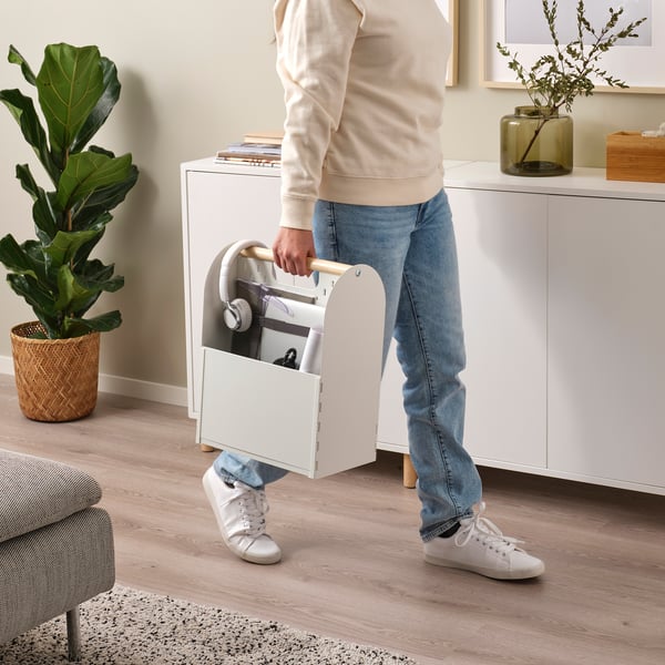 Person holding VATTENKAR portable organiser with laptop, headphones, and files, standing by white cabinets and plants.