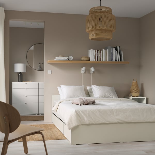 Minimalist bedroom with beige bed, white linens, wicker light, mirror, dresser, chair & rug.
