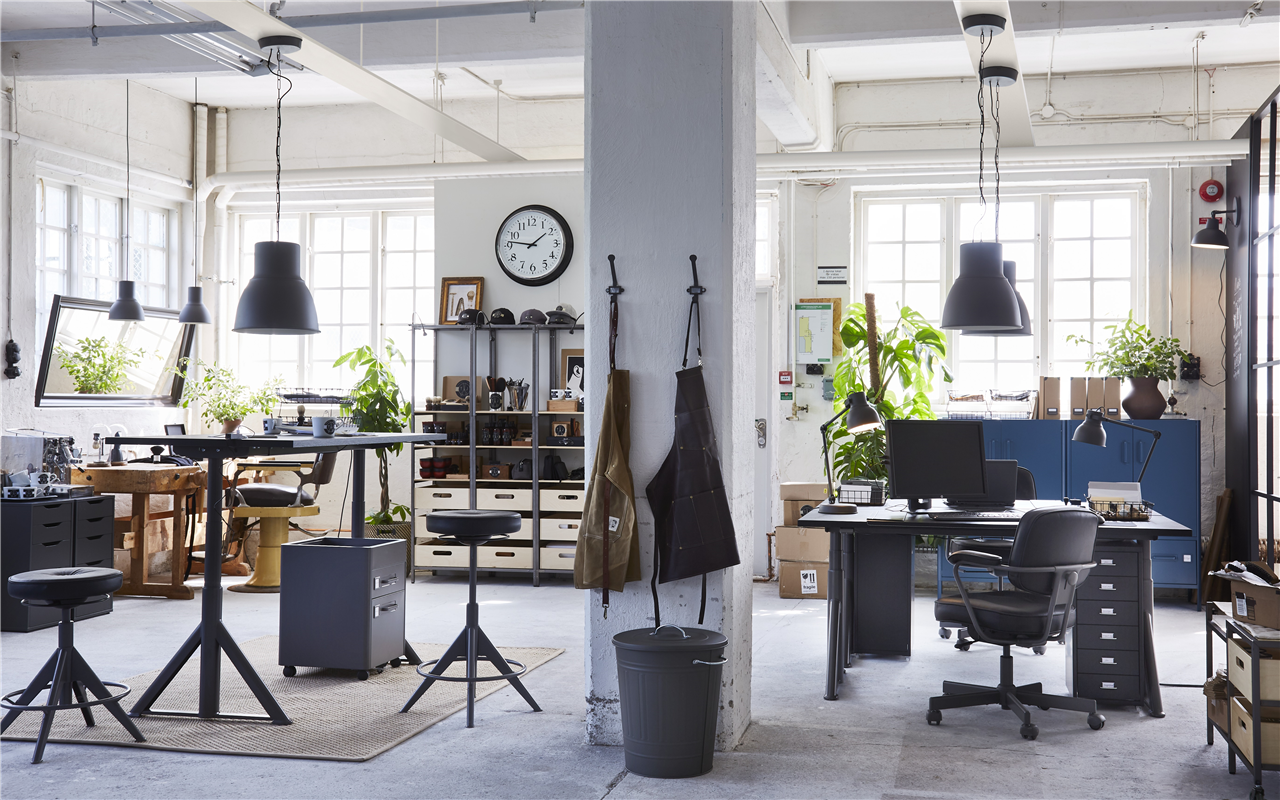 Industrial-style office space with shelves, workstations with IDÅSEN sit/stand desks and aprons hanging from a concrete pillar.