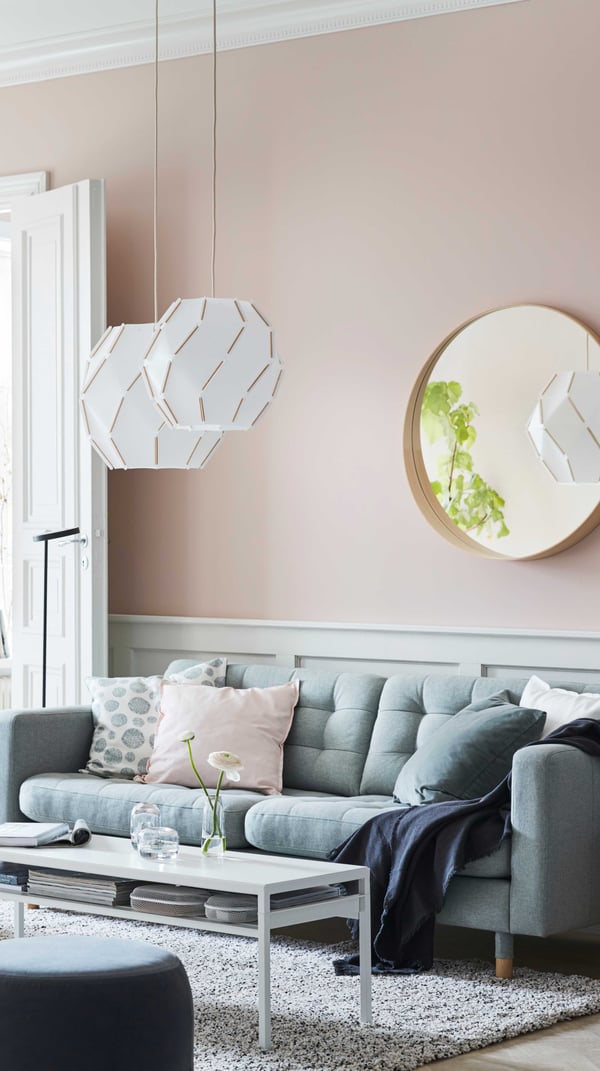 Ein hellrosa Wohnzimmer mit einem Spiegel an der Wand und Lampen, die von der Decke hängen. Ein hellgrünes Sofa mit Kissen, Überwurf und einem weissen Couchtisch mit Ablage vor dem Sofa.