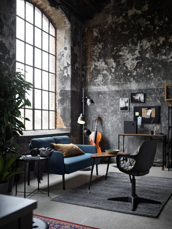 Es ist ein Raum im industriellen Flair. Es ist etwas dunkler eingerichtet. Auf der linken Seite sthet ein blaues Sofa und gegenüber ein schwarzer Stuhl auf einem grauen Teppich. Die Wände sind in einem weiß - Grau Muster gestrichen und an der Wand hängen 5 Bilder.