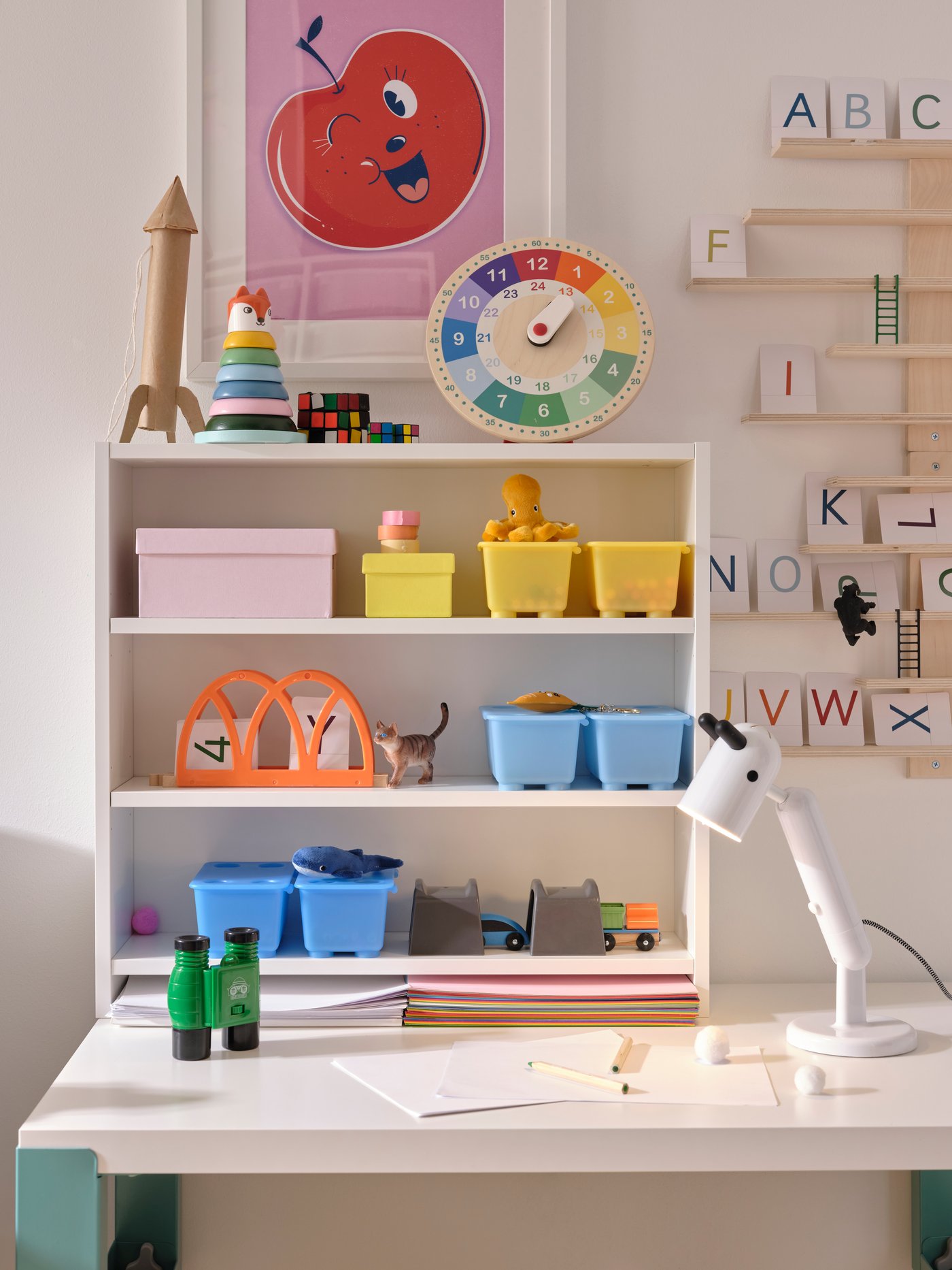 A white PÅHL desktop shelf on a desk holding blue, yellow and pink boxes with toys in. A KRUX LED work lamp is beside it.