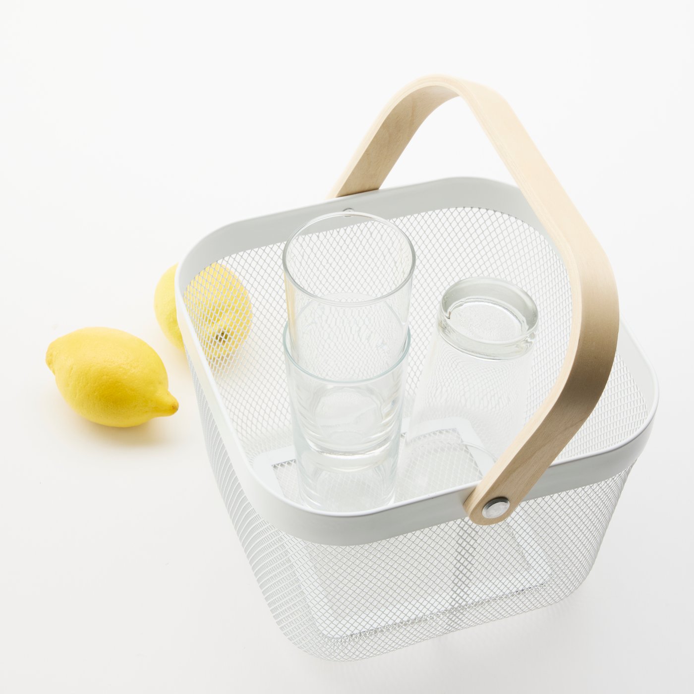 Close-up of a RISATORP mesh basket, containing a glass carafe and drinking glasses. Behind the basket, some lemons.