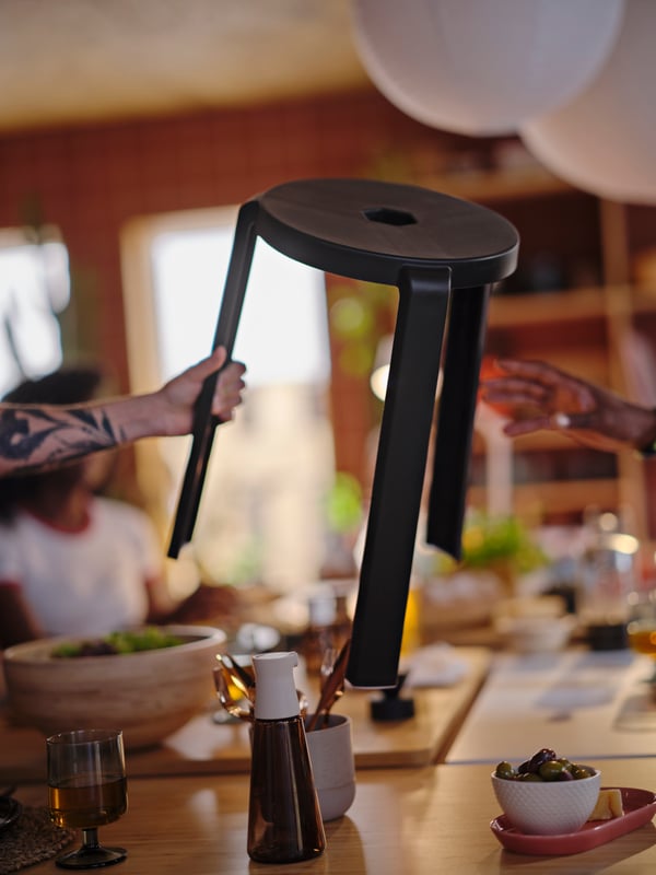 A tattooed arm is holding a dark grey IBBARBO stool in the air, reaching it over to a hand about to grab it.