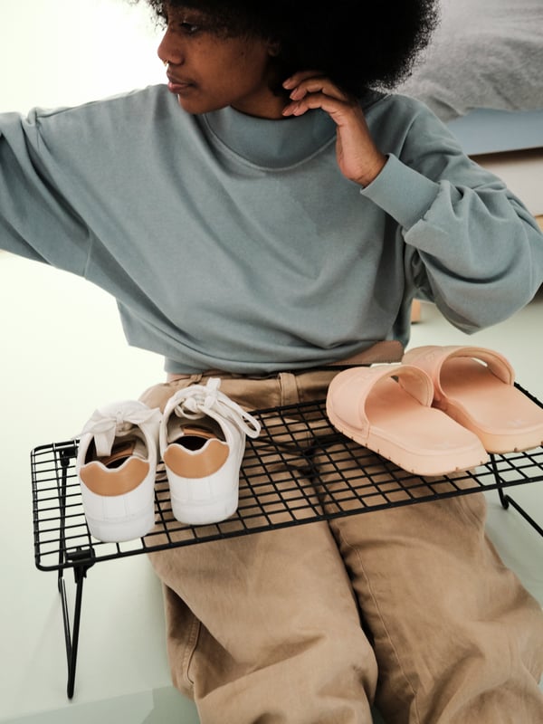 A young woman sitting on the floor while arranging two pairs of shoes on a black GREJIG shoe rack in steel.