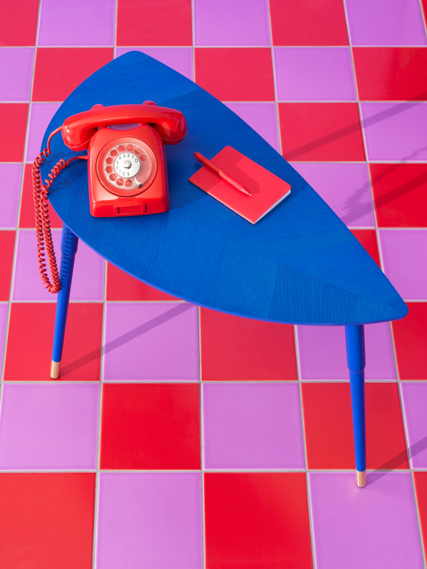 A red rotary dial telephone, notebook and pen are on a blue LÖVBACKEN side table on a red and lilac square tiled floor.