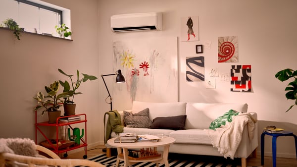t modernt vardagsrum med en vit soffa dekorerad med gröna och svartvita kuddar, en beige filt och ett litet runt träbord med böcker och en kopp. Till vänster står en röd vagn med gröna växter, och ovanför soffan hänger abstrakta konstverk i olika färger. En svart golvlampa står bredvid soffan, och en vit luftkonditioneringsenhet är monterad på väggen.