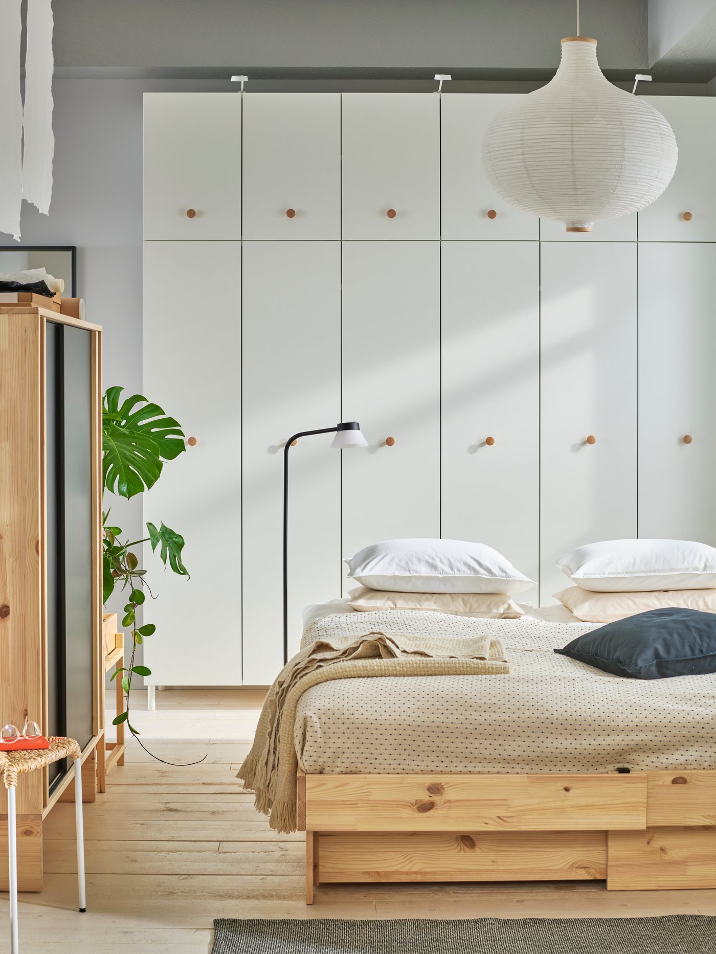 Chambre à coucher dans des tons beige et blanc, avec un lit en bois naturel et une literie beige. Au fond, une rangée d'armoires PLATSA blanches.